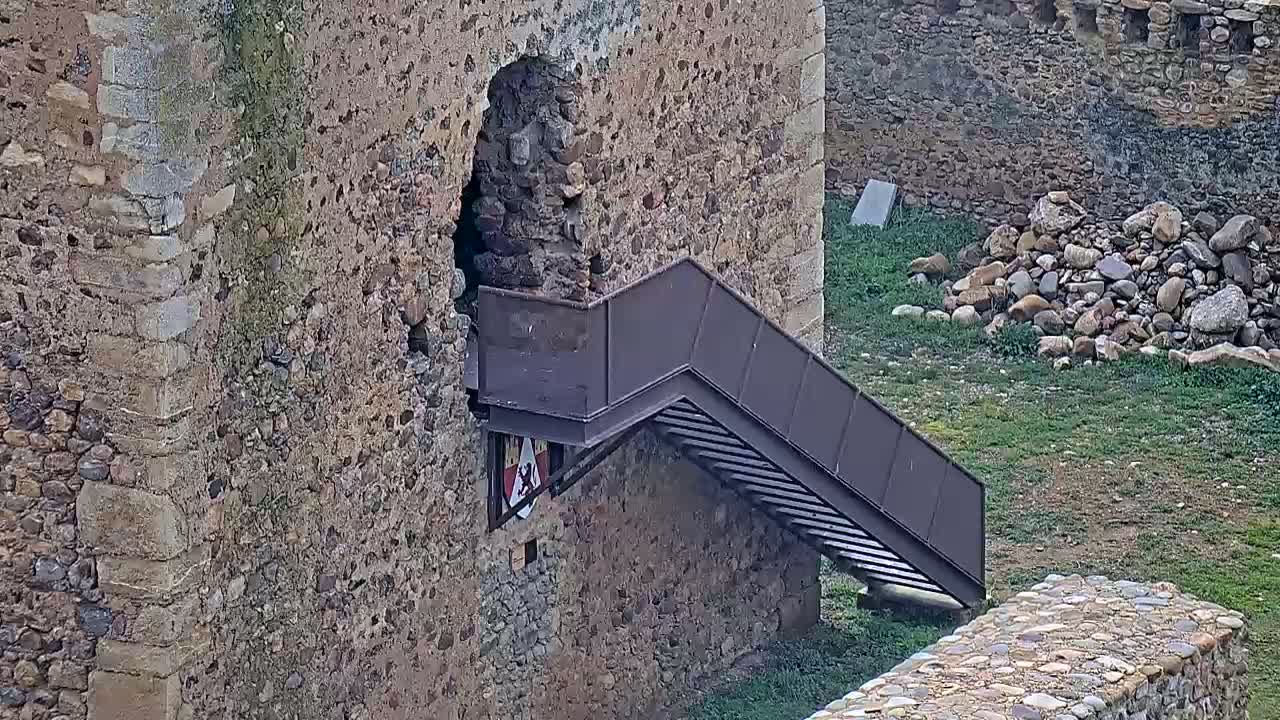 A dark brown metal staircase extends from a stone wall, leading down to a lower section of a stone structure with overgrown grass.
