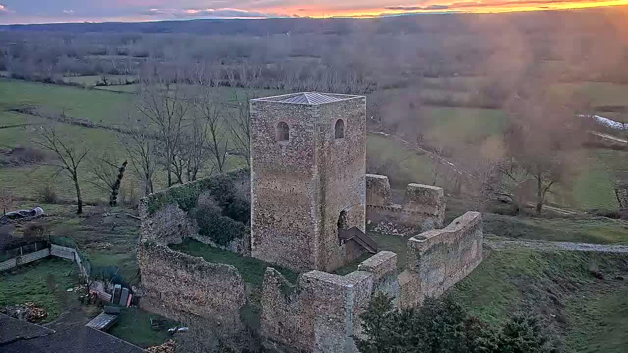 An aerial view shows the stone ruins of a castle or tower, partially intact, set against a landscape of farmland under a sunset sky.