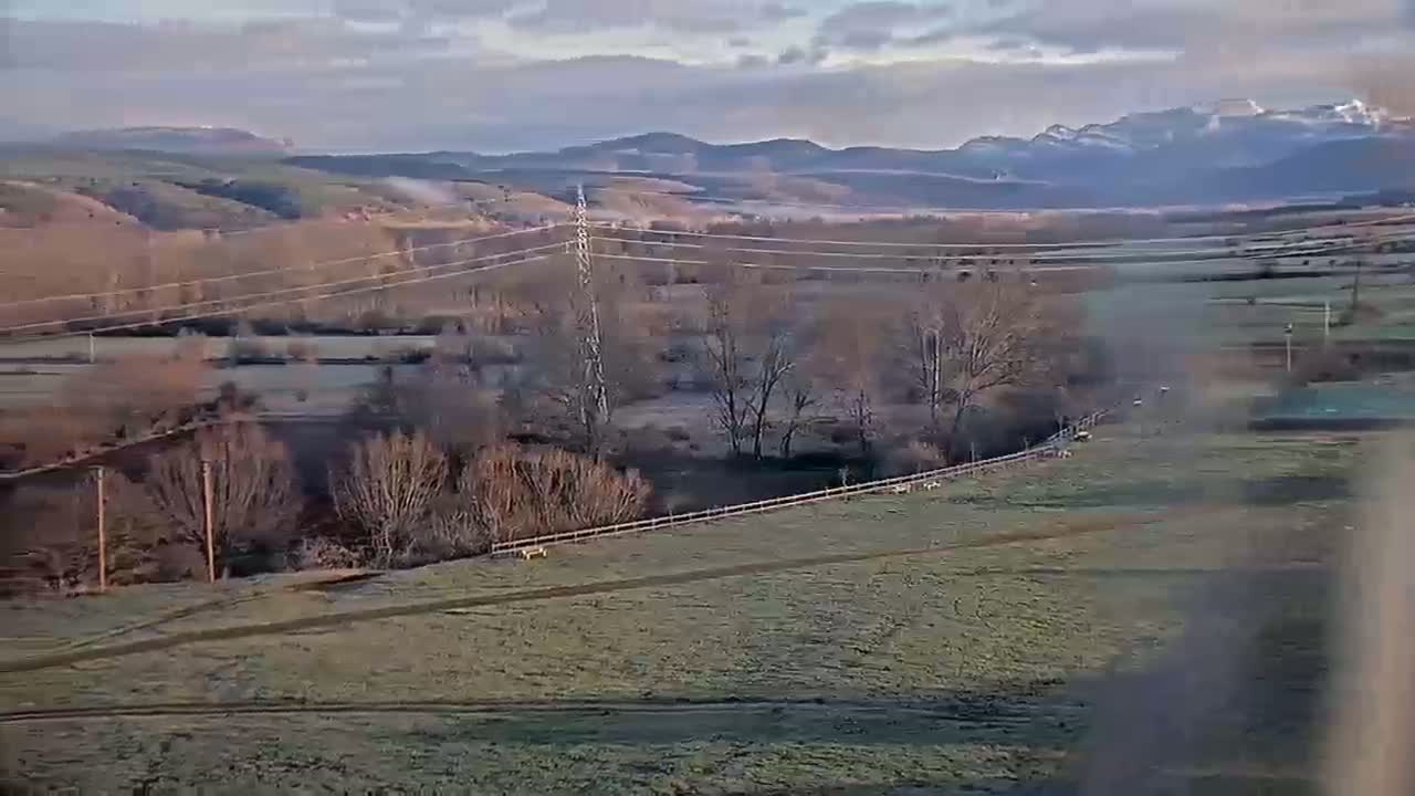 A high-angle view of a pastoral landscape under a partly cloudy sky shows a valley with bare trees, a river, and a field with some animals and fences, all set against a backdrop of distant mountains.