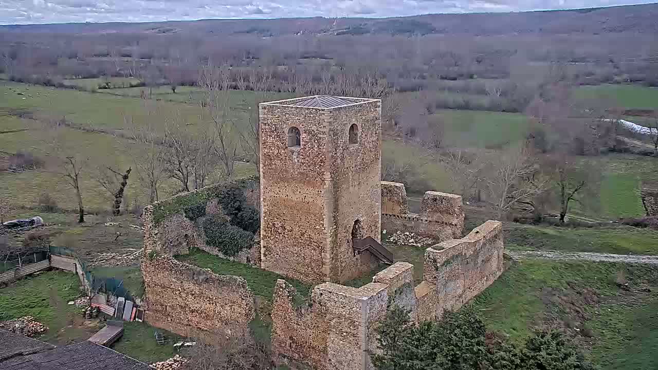 An aerial view shows the stone ruins of a tower and surrounding walls, situated in a rural landscape under a cloudy sky.