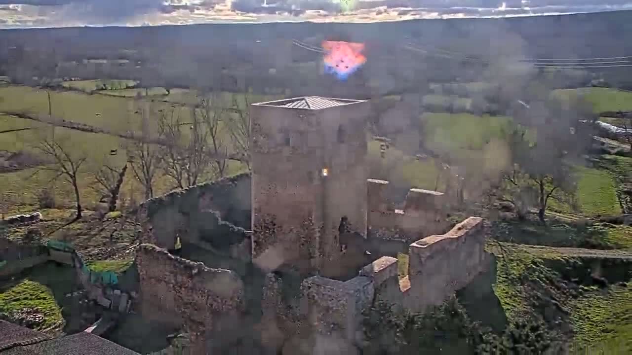 An aerial view shows the partially ruined stone walls of a castle or tower on a partly sunny day in a rural landscape with green fields and bare trees.