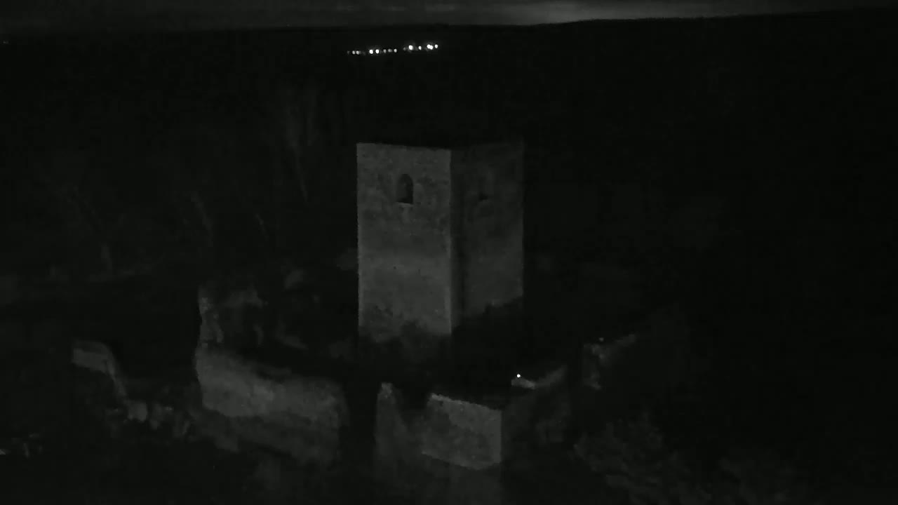 A dark, nighttime view shows a stone tower and surrounding structures faintly illuminated against a very dark background.