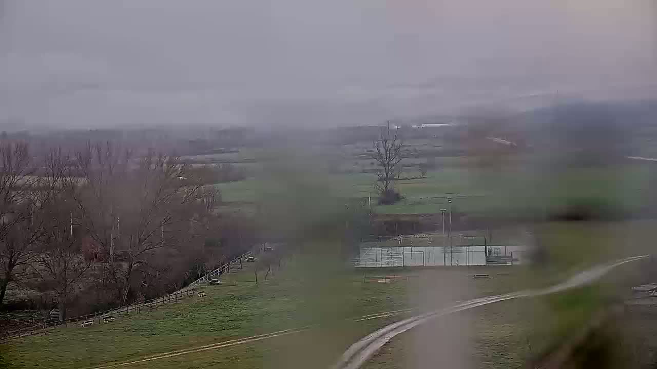 A hazy, overcast day reveals a view of a green field with a tennis court, bare trees, and a dirt path, all seen from a distance.