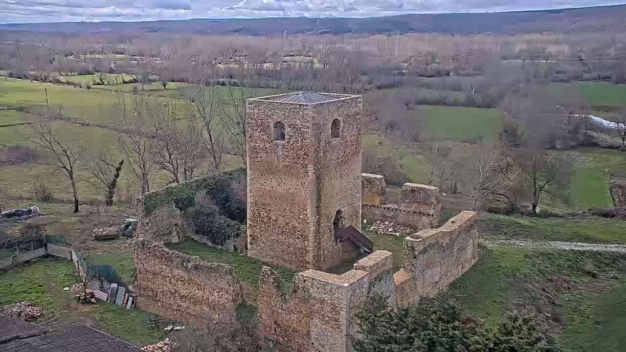 A partly ruined stone tower and wall stand amidst a landscape of bare trees and green fields under a cloudy sky.