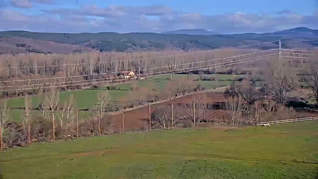 A partly cloudy day reveals a rural landscape of fields, bare trees, and a single house, all under power lines stretching across a valley towards distant mountains.