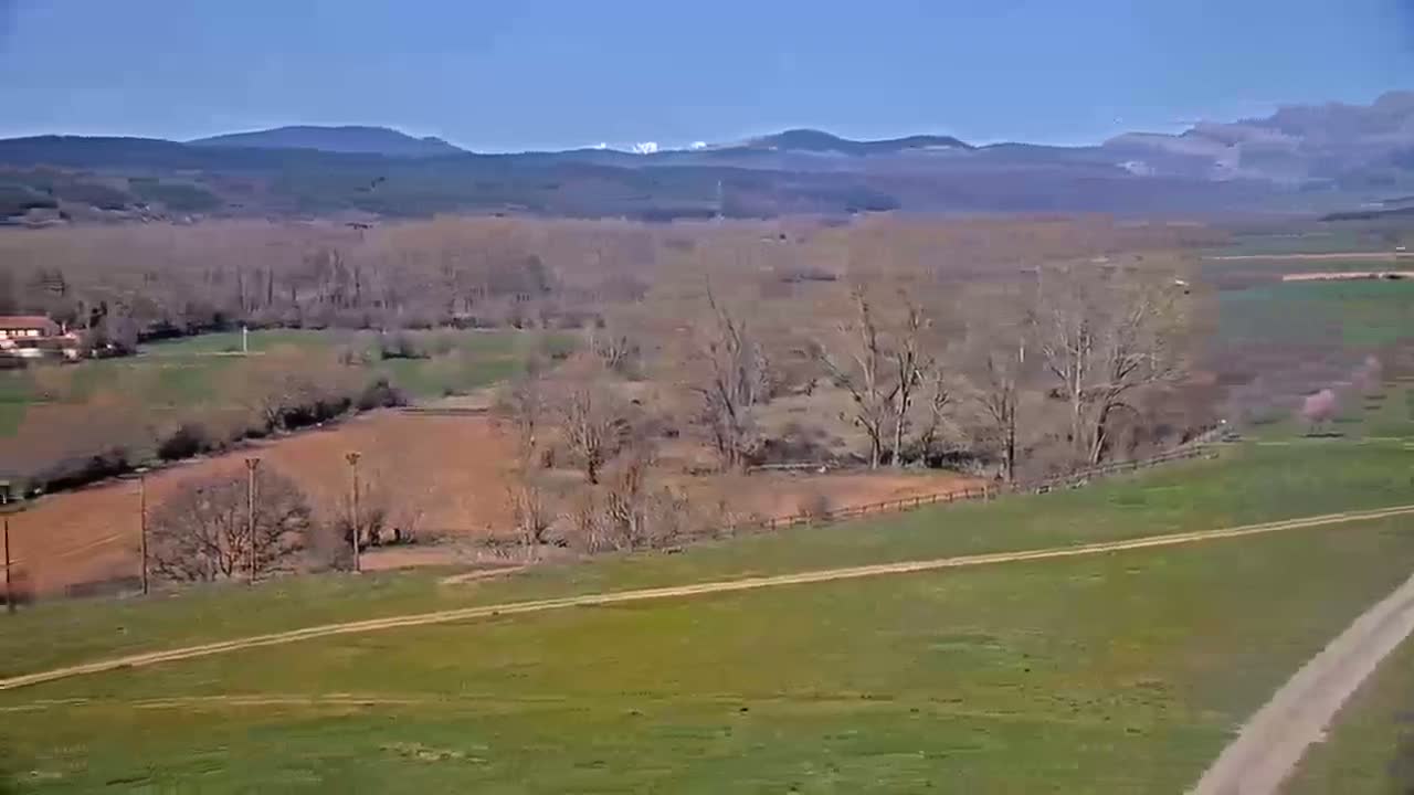 A high-angle view shows a rural landscape under a clear blue sky, featuring bare trees, brown and green fields, a dirt road, and distant mountains with snow-capped peaks.