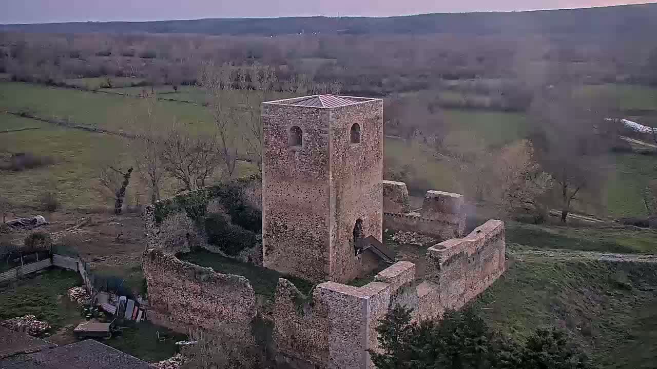 An aerial view shows the ruins of a stone tower and surrounding walls, situated on a grassy hillside under a dusky sky.