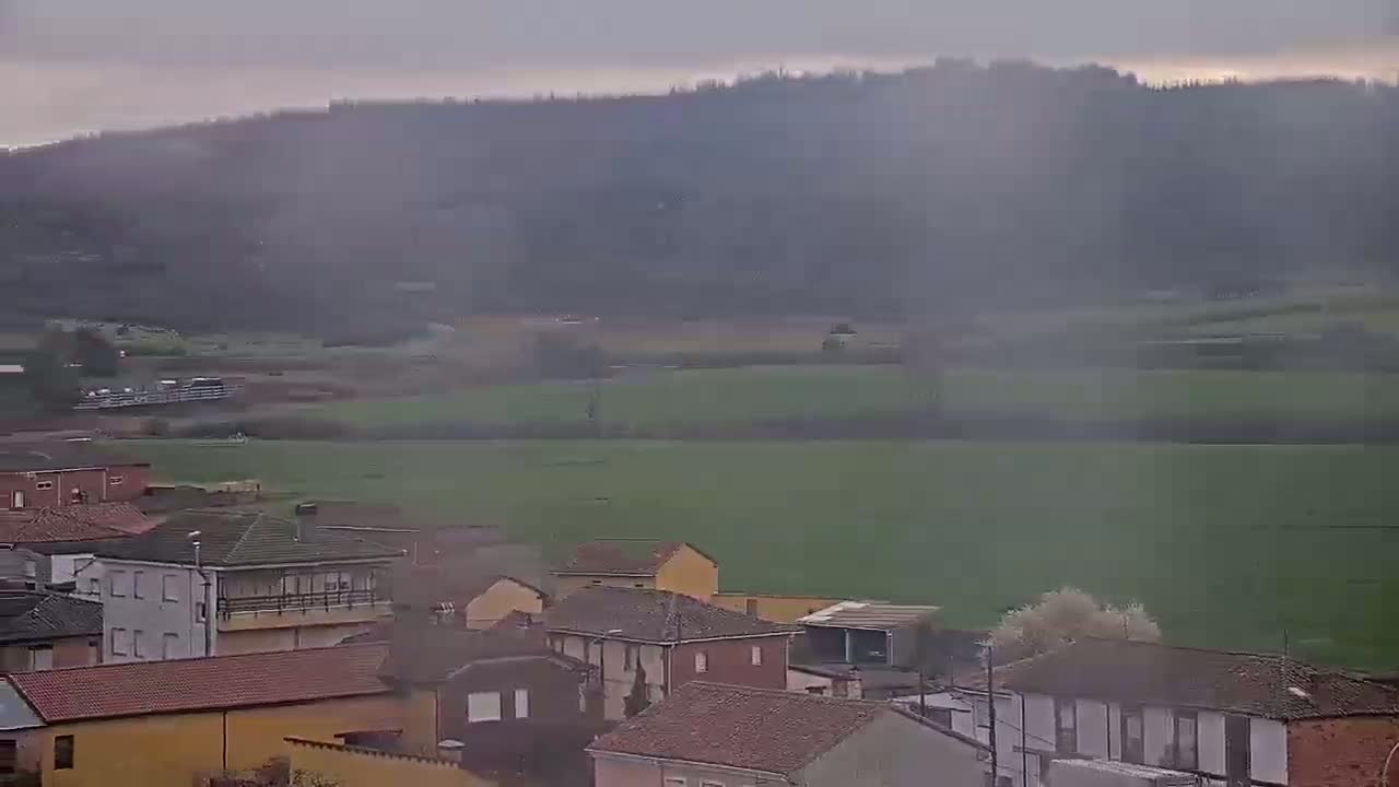 A hazy view shows a small village nestled in a valley, with a vast green field extending towards a misty hillside.