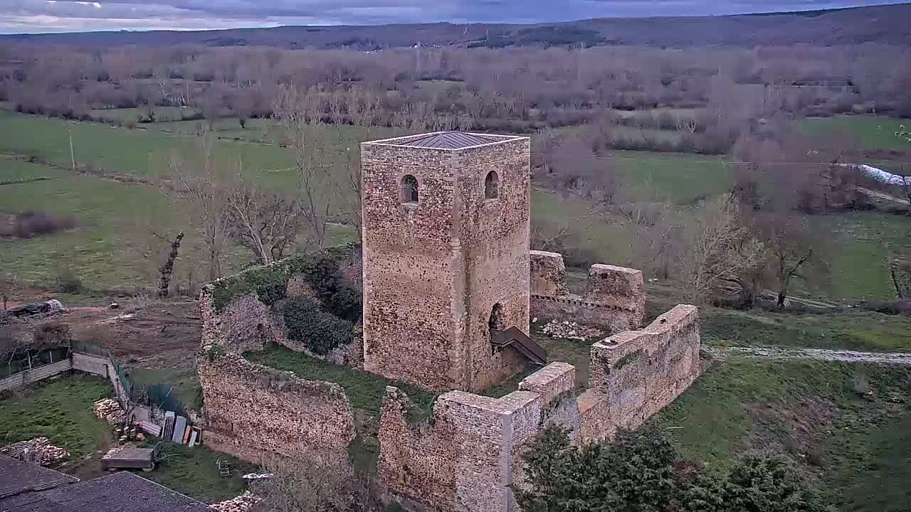 An aerial view shows the ruins of a stone tower and surrounding walls, set in a rural landscape under a cloudy sky.