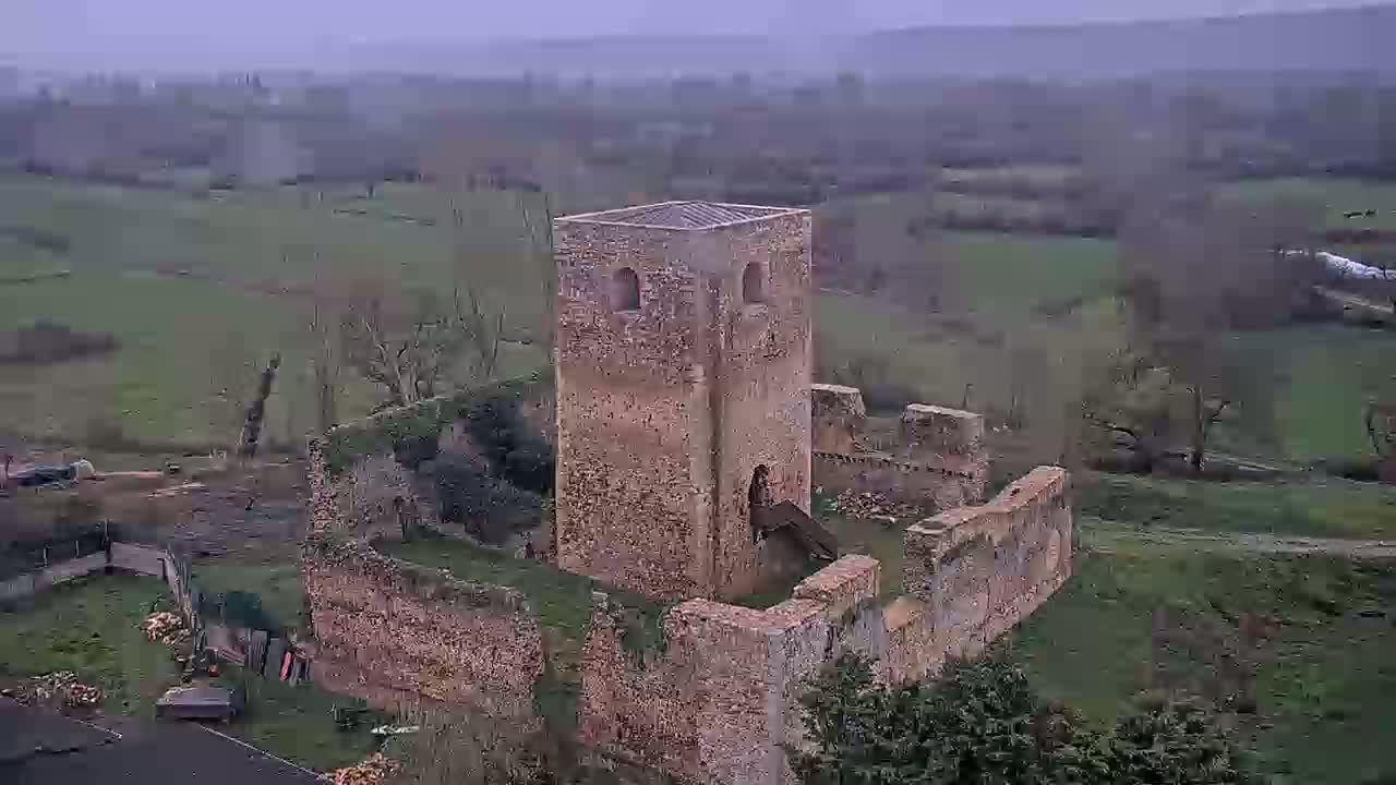 An aerial view shows the ruins of a stone tower and surrounding walls, set in a verdant landscape on an overcast day.