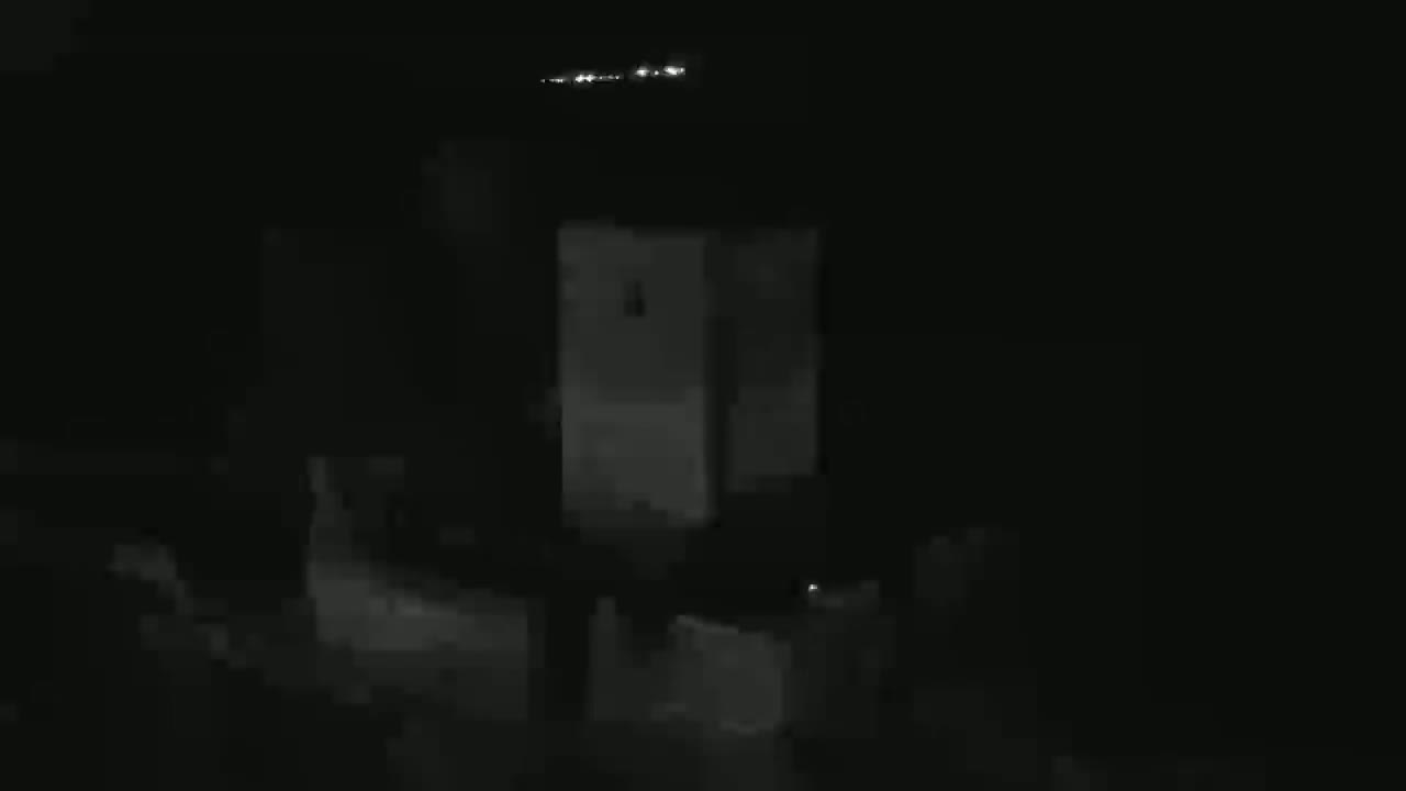 A dimly lit, rectangular structure is visible at night, with faint lights in the distance.