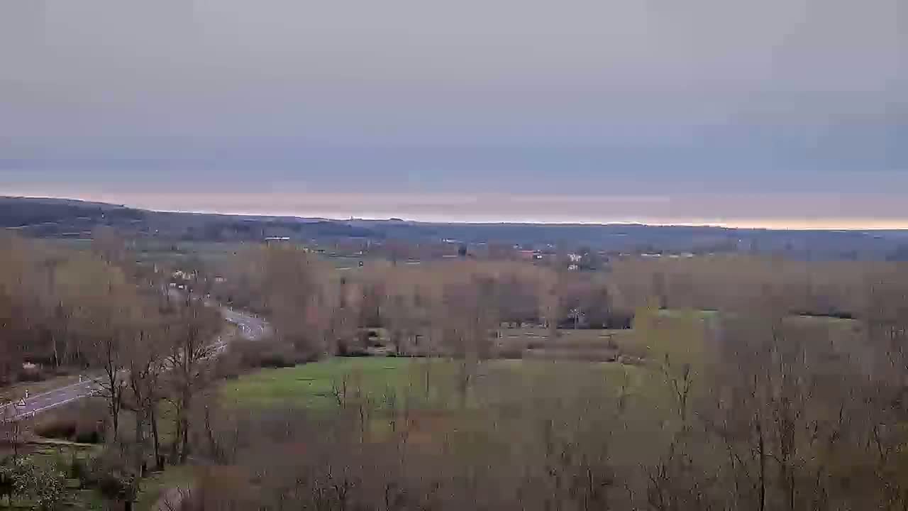 A mostly cloudy day reveals a vast, flat landscape of leafless trees, a paved road, and patches of green fields stretching to distant hills.