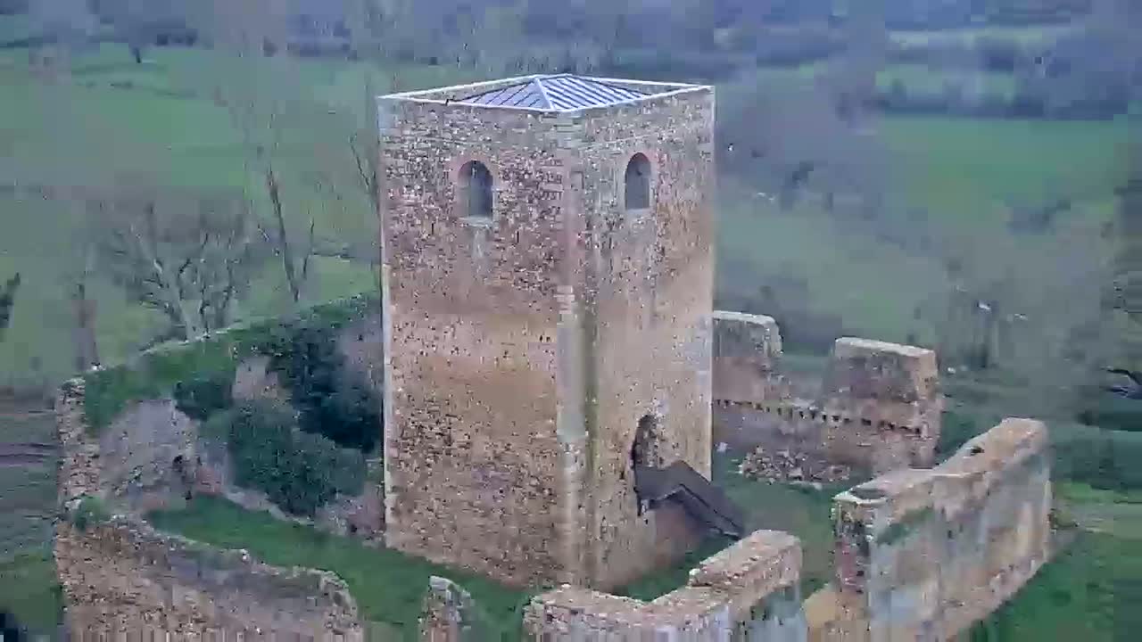 An aerial view shows the stone ruins of a tower and surrounding walls, overgrown with grass and vegetation, on an overcast day.