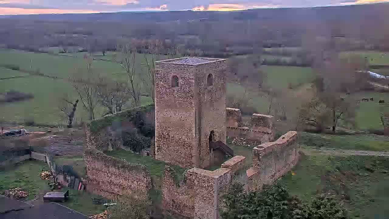 An aerial view shows the ruins of a stone tower and castle walls on a grassy hillside under a partly cloudy sky.