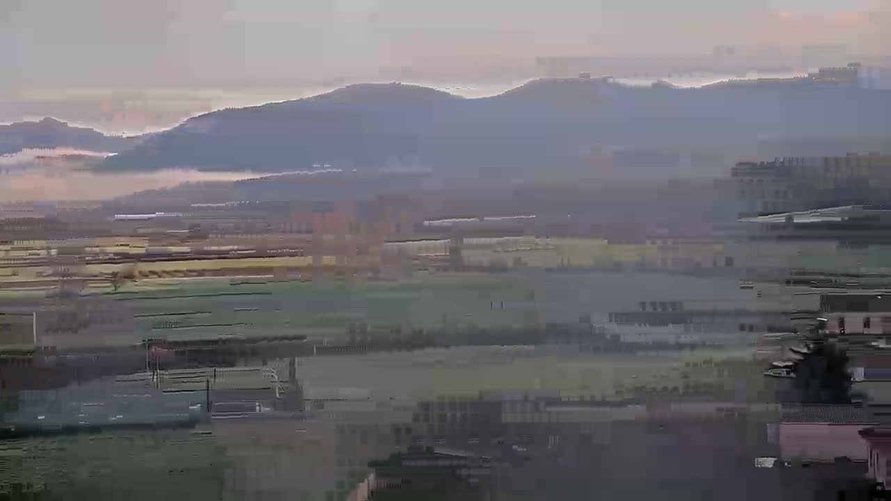 A hazy, possibly foggy, landscape shows distant mountains, a valley of fields and buildings, and a foreground of buildings and trees.