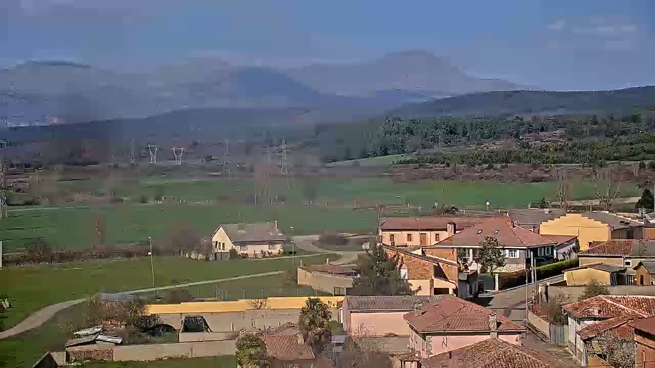 A small village with red-tiled roofs sits in a valley under a clear sky, with mountains and green fields visible in the distance.