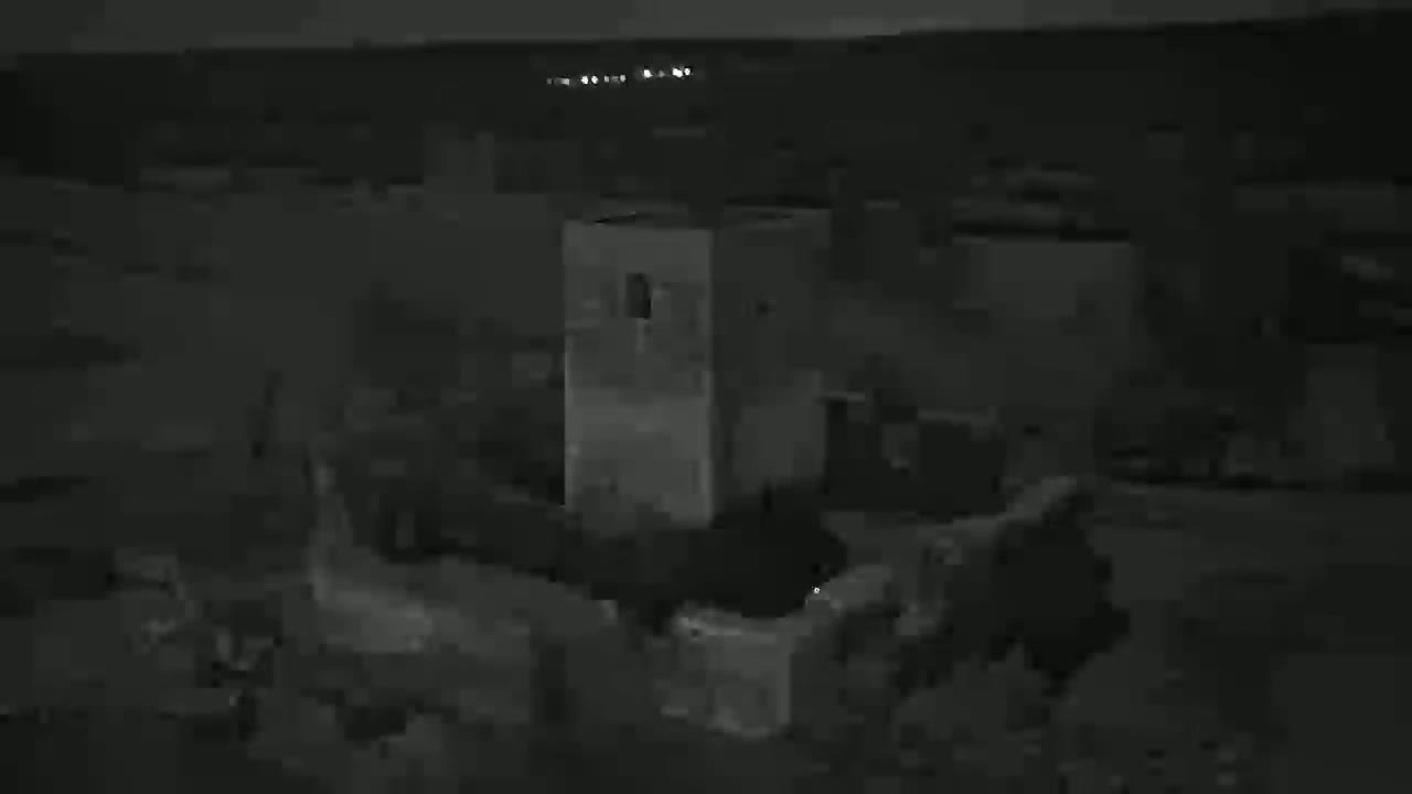 A nighttime, low-light image shows a tall, light-colored rectangular structure among other dark, indistinct structures, with a few distant lights visible in the background.