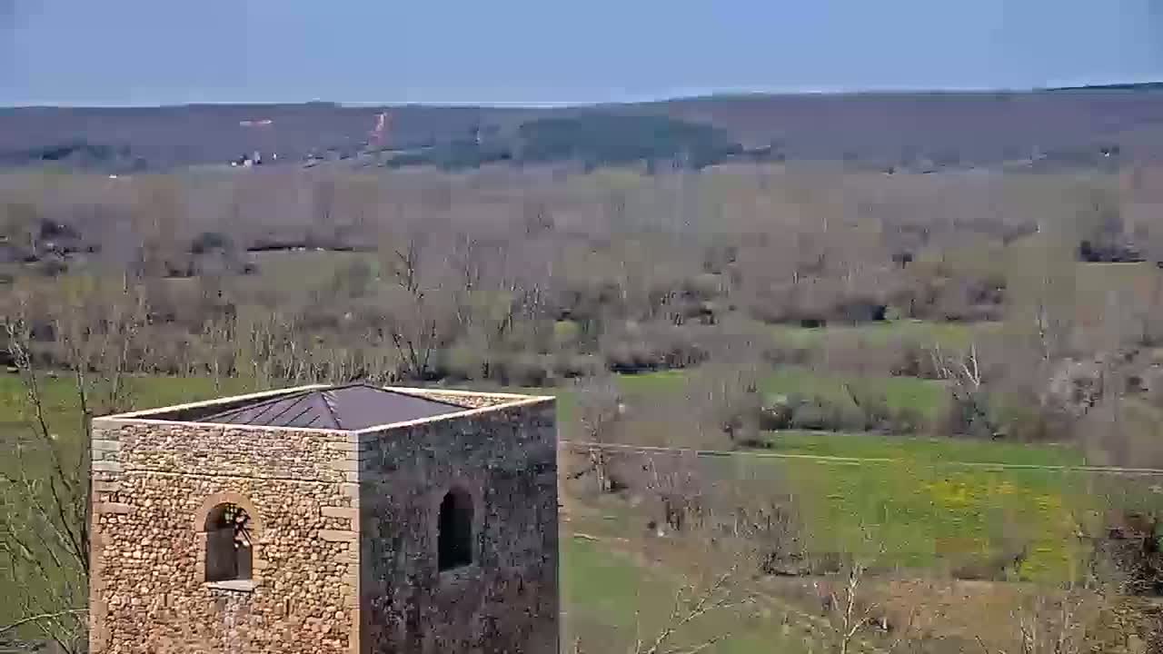 A stone tower with a flat roof overlooks a valley of bare trees and grassy fields under a clear blue sky.