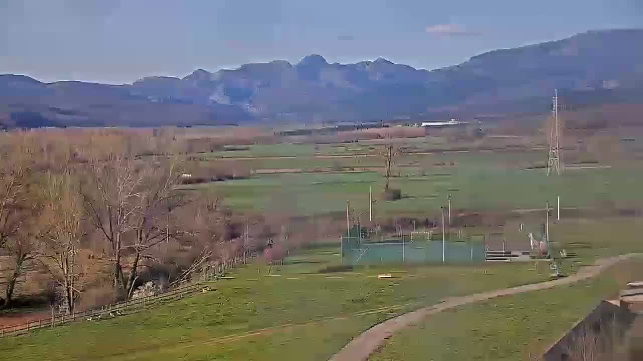A mostly sunny view of a rural landscape with bare trees, green fields, a distant mountain range, and a small fenced-in area.