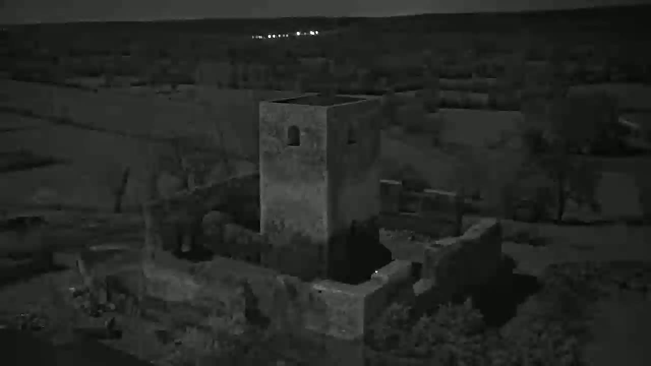 An aerial nighttime view shows the ruins of a stone structure, possibly a tower or castle, surrounded by dark vegetation under a clear, dark sky.
