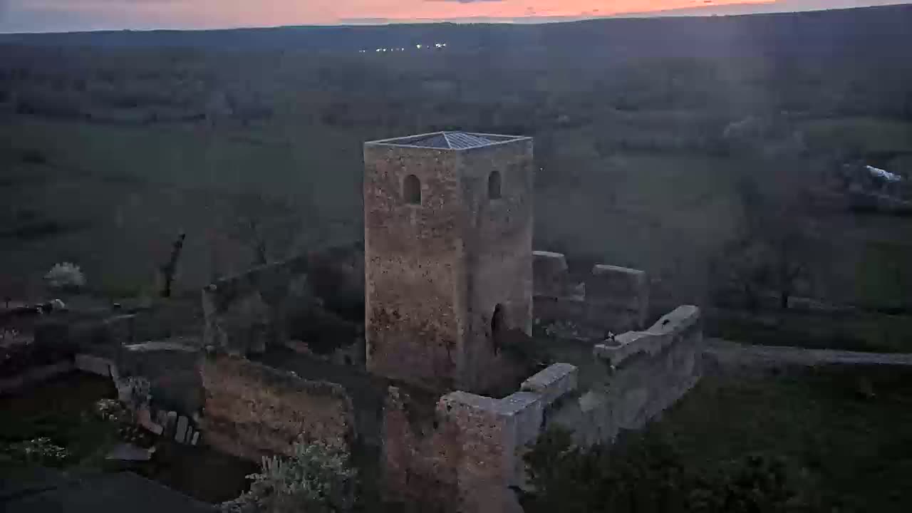 An aerial view shows the ruins of a stone tower and surrounding walls at dusk, under a pinkish-orange sunset.