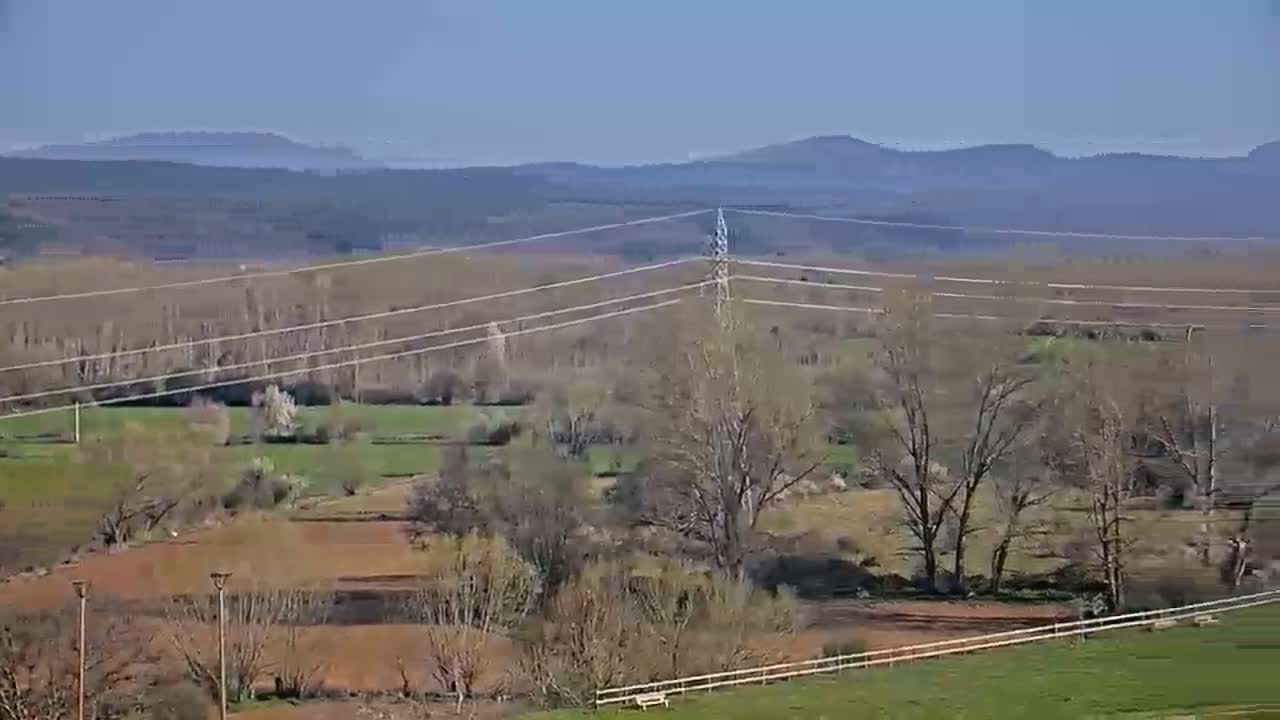 A long-range view shows a rural landscape under a clear blue sky, featuring bare trees, fields of brown earth and green grass, power lines, and distant mountains.