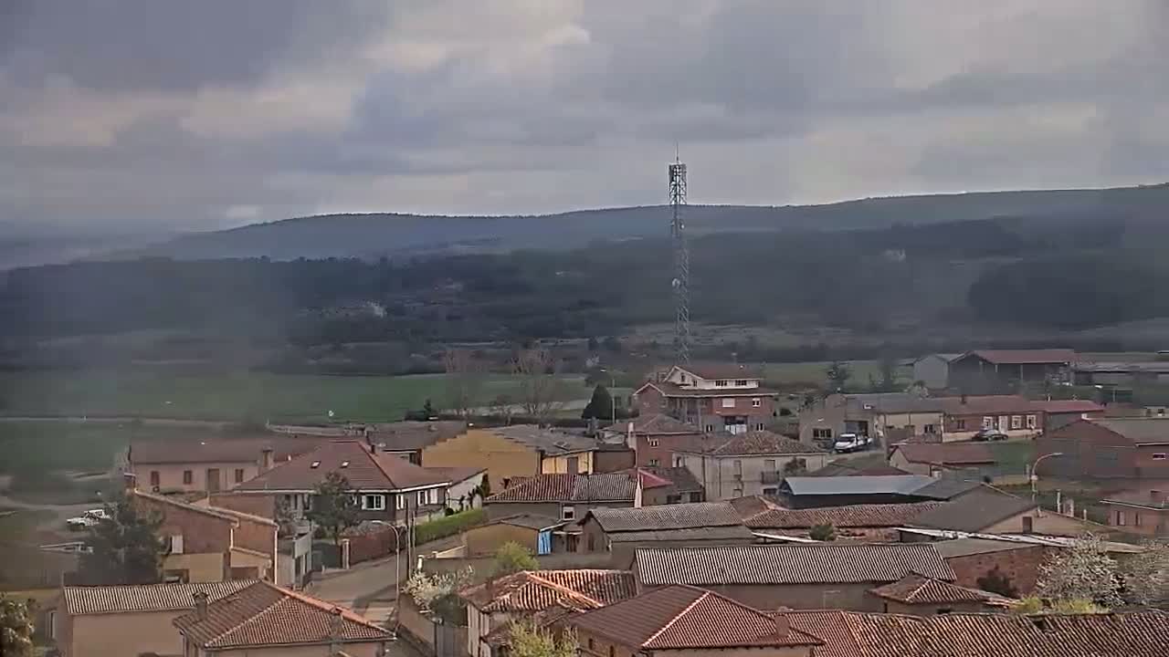 A small town of brown-roofed buildings sits nestled in a valley under a cloudy sky, with a tall communication tower visible in the middle ground.