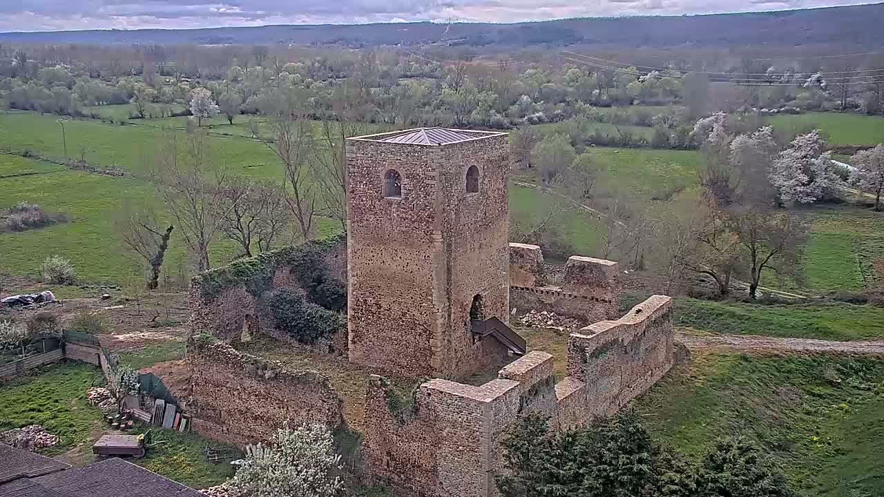 Stone ruins of a tower and surrounding walls sit on a grassy hillside overlooking a verdant valley under a cloudy sky.
