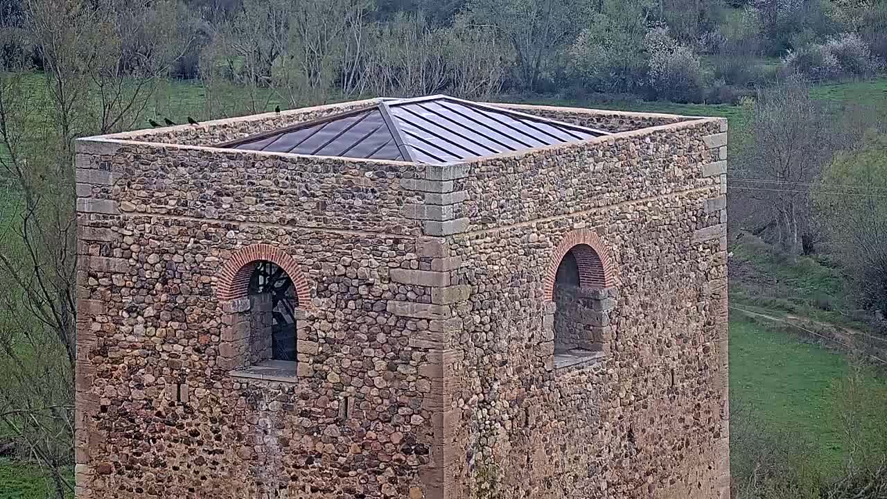 A stone tower with a flat, metal roof sits atop a grassy hillside, several birds perched on its edges.