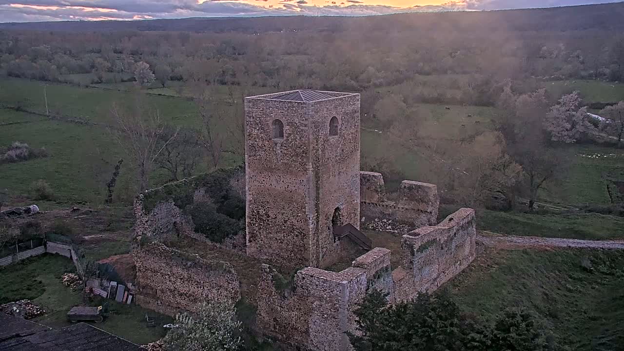 An aerial view shows the stone ruins of a castle tower and surrounding walls overlooking a valley at sunset.