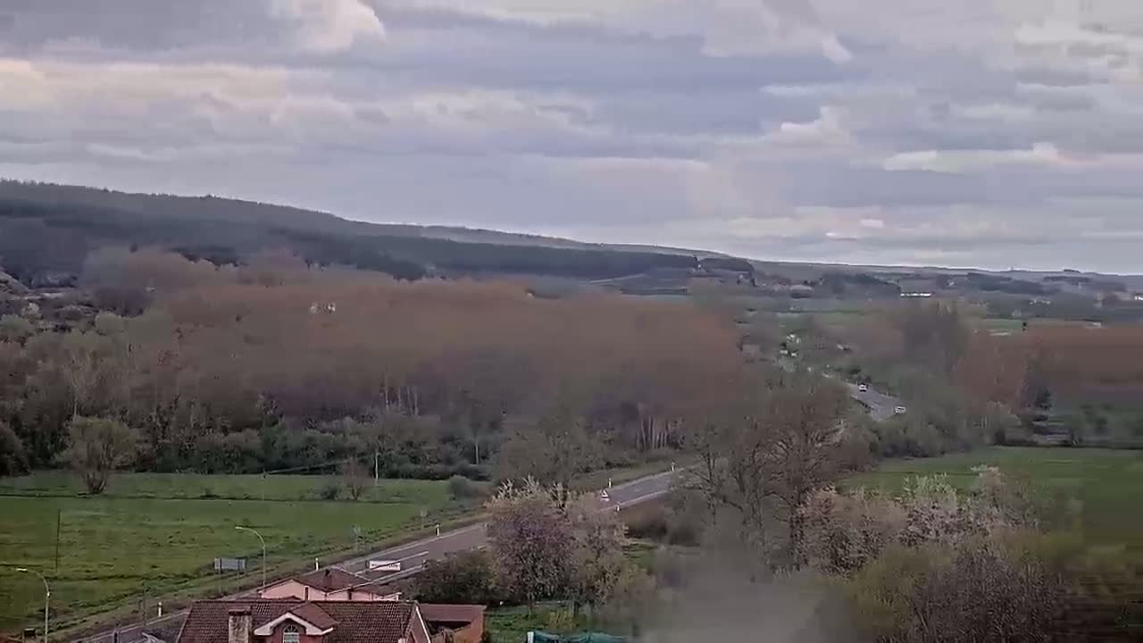 A long road cuts through a rural landscape of fields and bare trees under a cloudy sky.