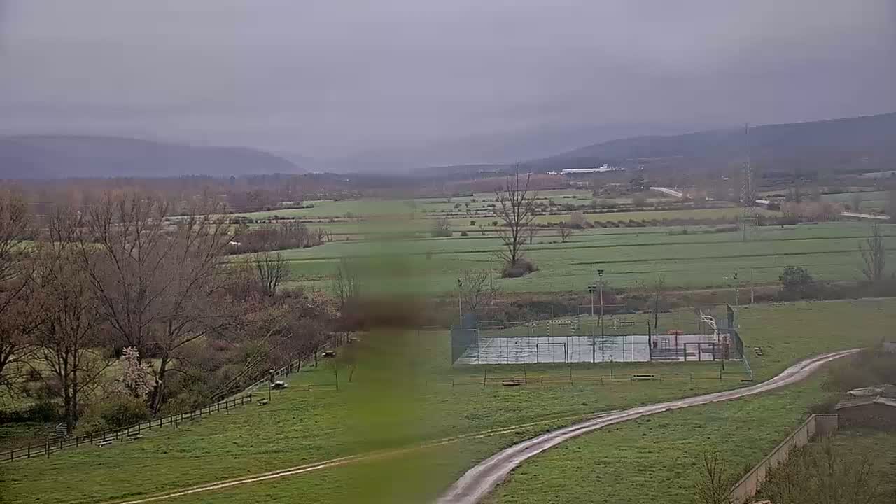 A panoramic view of a valley on an overcast day shows a network of fields, bare trees, and a paved court.