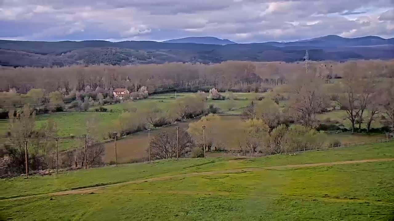 Under a cloudy sky, a sprawling green valley dotted with trees, a few houses, and a dirt road stretches towards distant, hazy mountains.