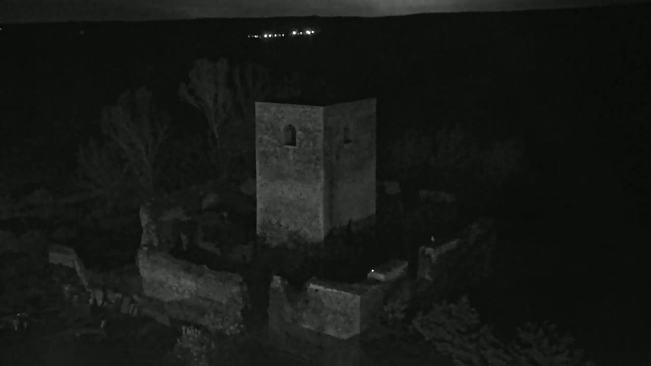 An aerial nighttime view shows the stone ruins of a structure, possibly a tower, surrounded by dark vegetation under a mostly clear sky.