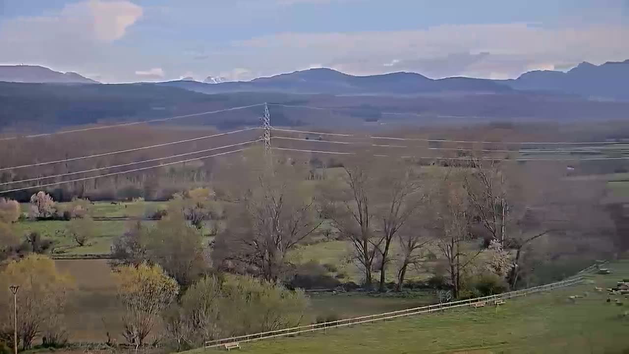 A pastoral landscape under a partly cloudy sky features power lines stretching across a valley of bare trees, green fields, and distant mountains.