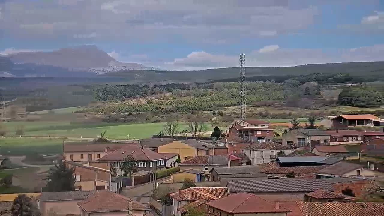 A small town with red-tiled roofs nestles in a valley under a cloudy sky, with a mountain visible in the distance.