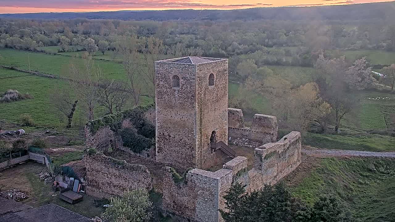 An aerial view shows the ruins of a stone tower and castle walls at sunset overlooking a verdant valley.
