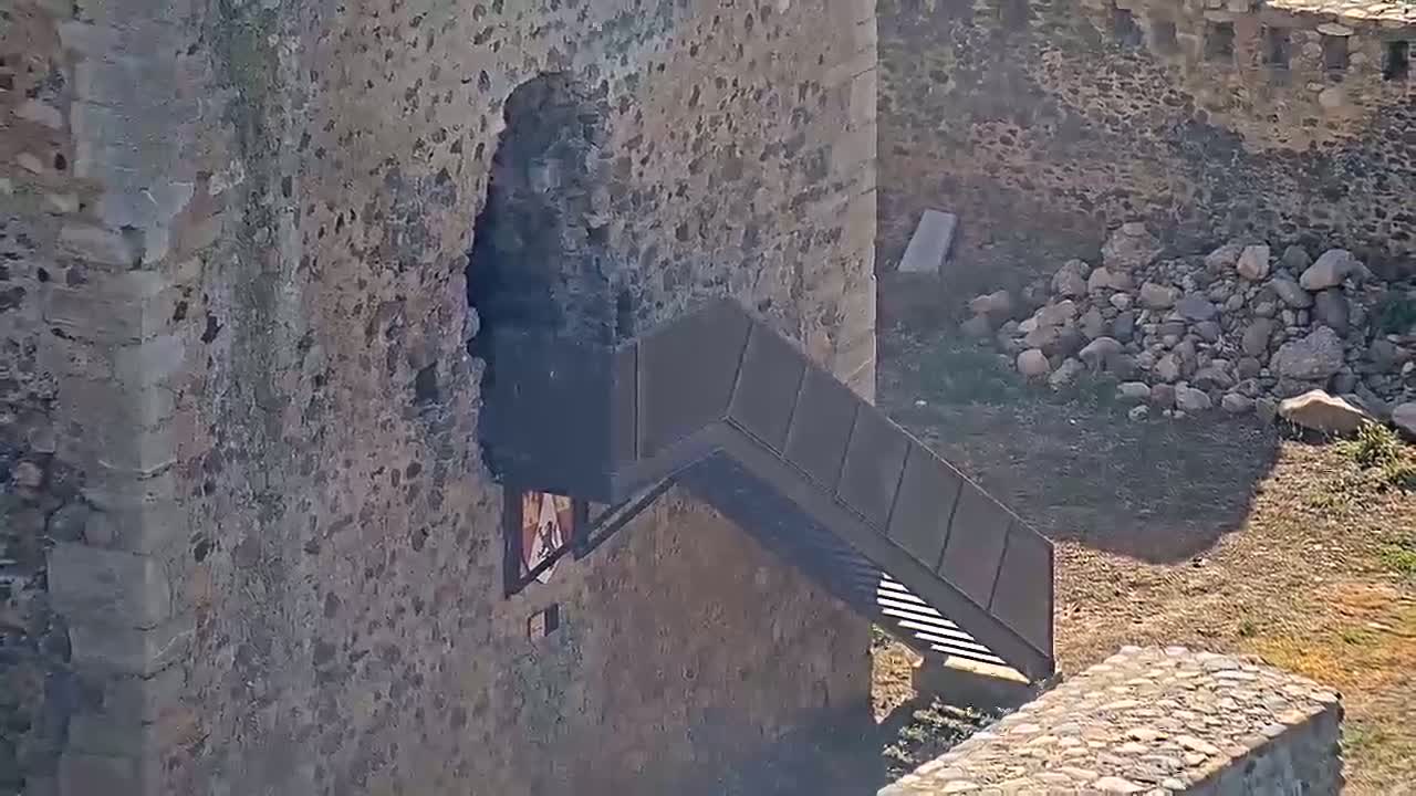 A modern metal staircase extends from a stone castle wall,  on a sunny day, towards a pile of rocks and dry ground.
