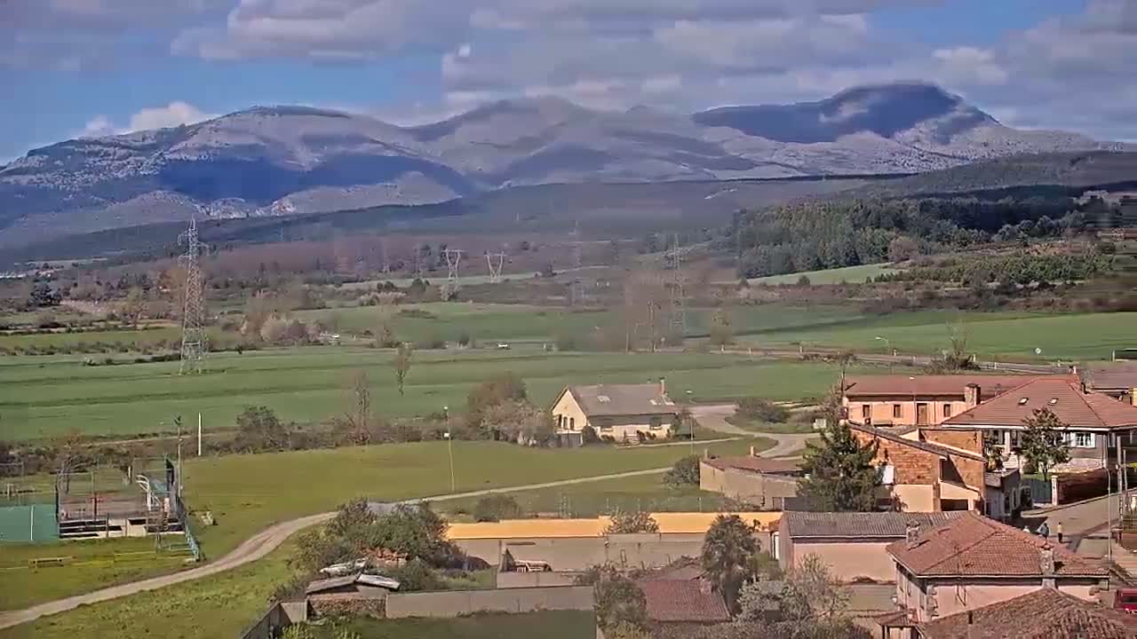 A partly cloudy day reveals a sprawling rural landscape dotted with buildings, fields, and power lines, all set against a backdrop of snow-capped mountains.