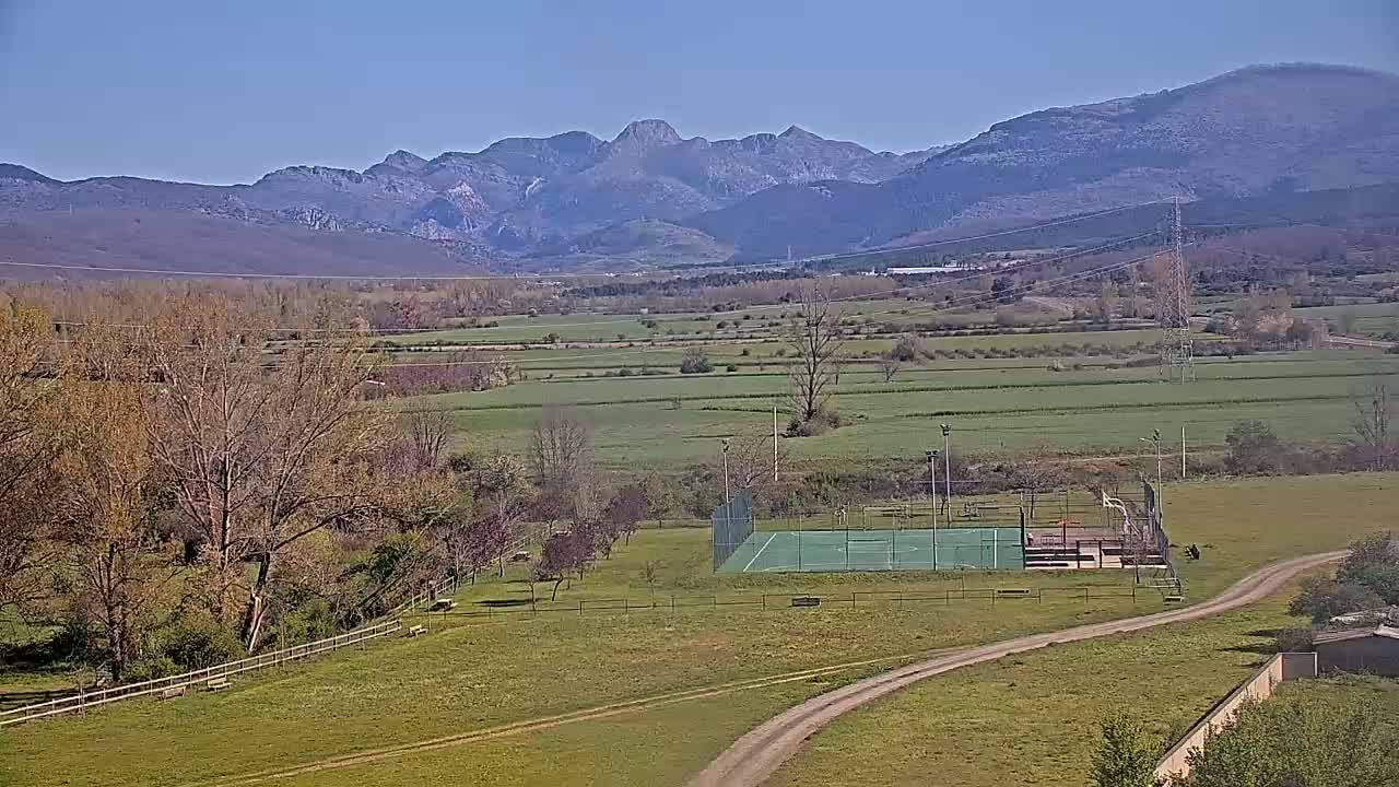 A green outdoor sports court sits in a grassy field, with bare trees and a distant mountain range visible under a clear blue sky.