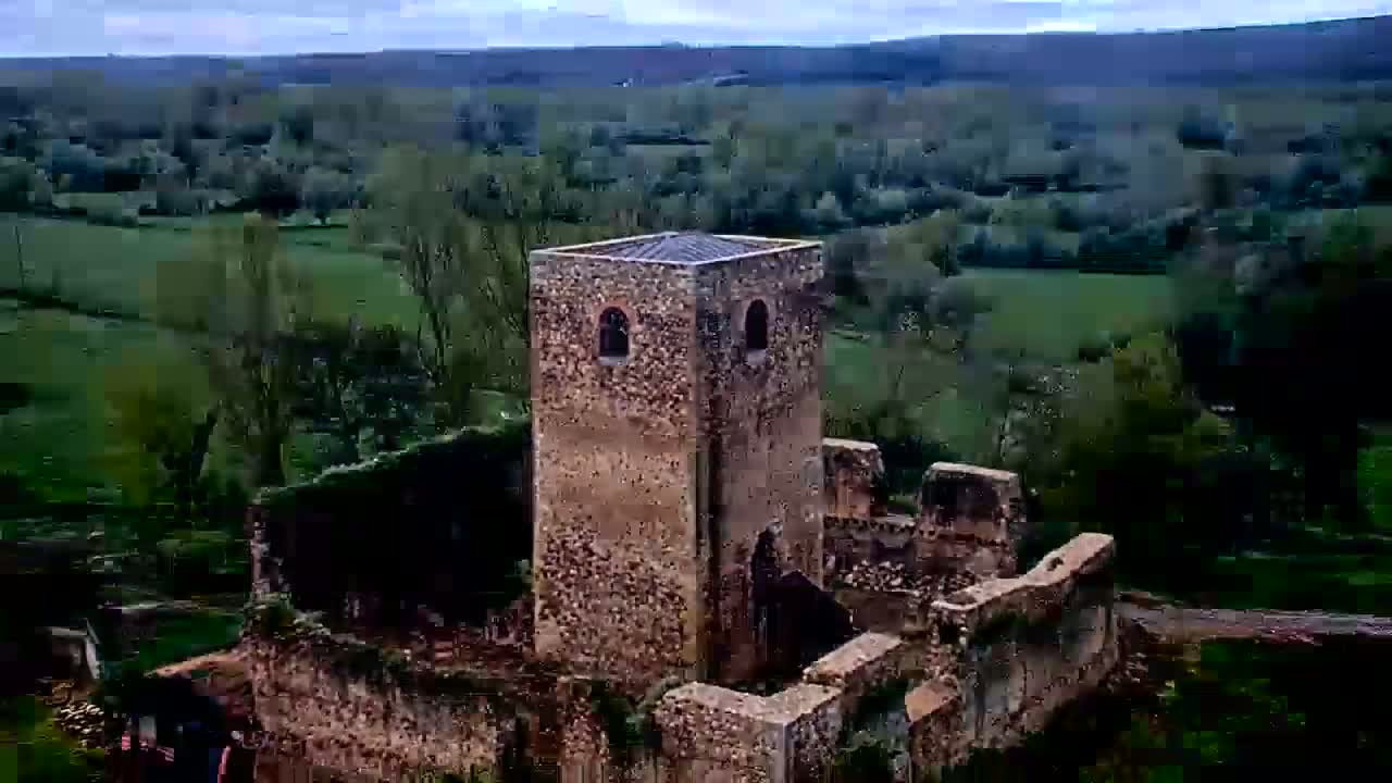 An aerial view shows the stone ruins of a castle, including a standing tower, overlooking a verdant landscape under a cloudy sky.