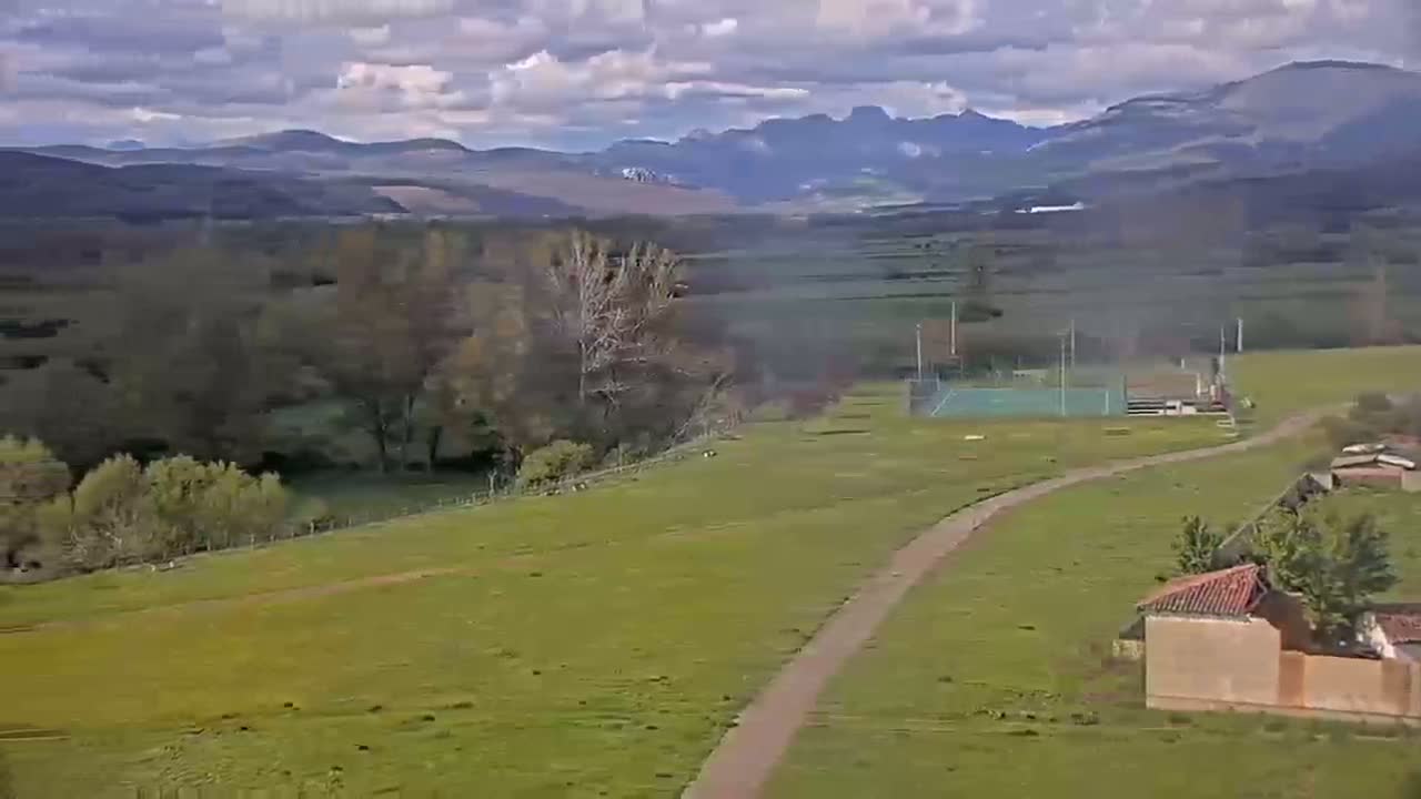A sprawling green field with a winding path leads to a small sports court, nestled beneath a partly cloudy sky and distant mountains.