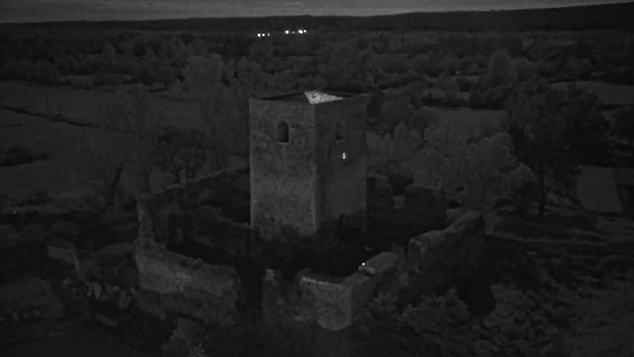 An aerial, nighttime view shows a stone tower and surrounding ruins nestled amongst dark trees under a twilight sky.