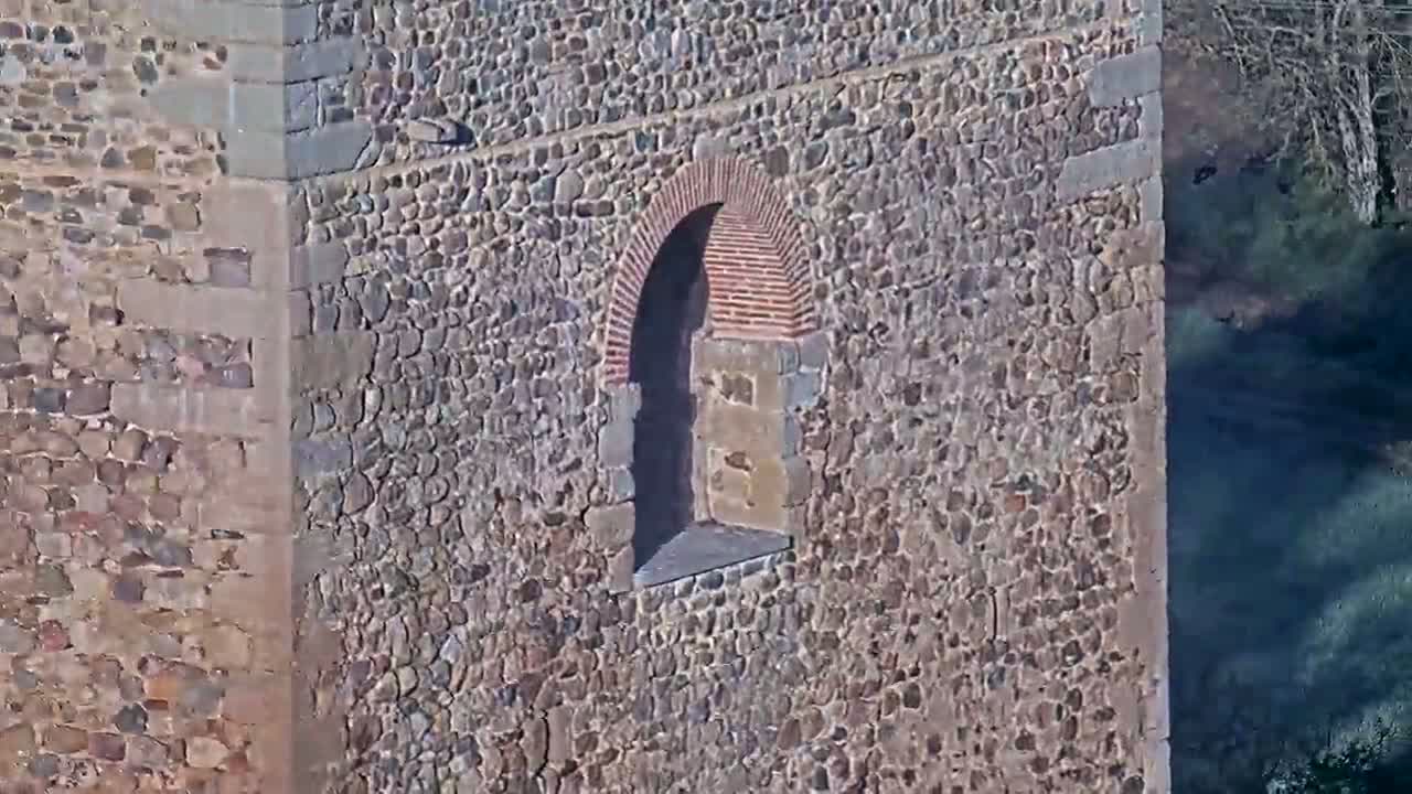 A stone wall with a small arched window, partially made of brick, is seen next to a shadowy area of what seems to be a wooded hillside.