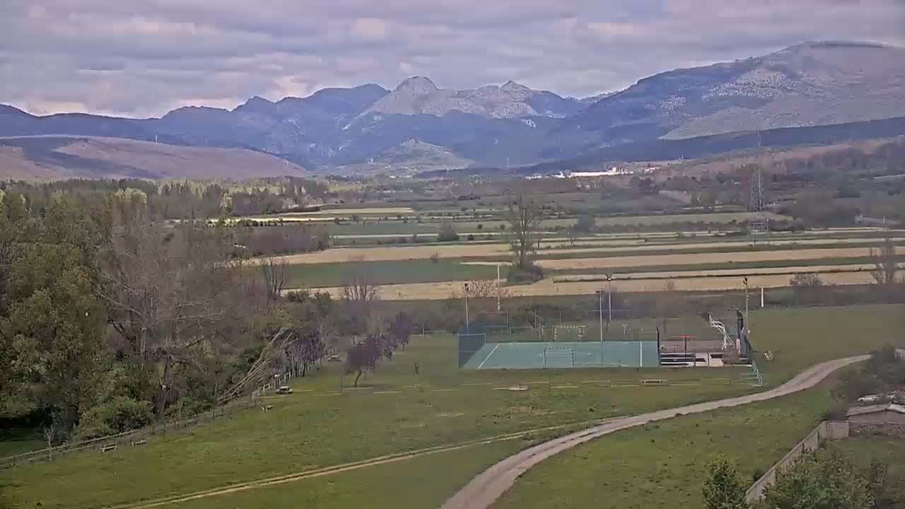 A sprawling landscape under a cloudy sky features a valley of farmland and a sports court, all set against a backdrop of distant mountains.