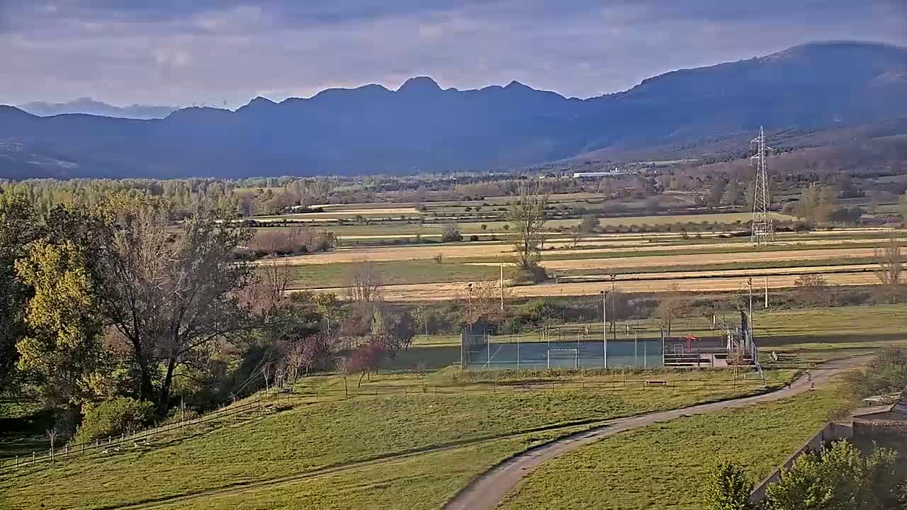 A paved path winds through a grassy field towards a tennis court, with a valley of farmland and distant mountains under a partly cloudy sky.