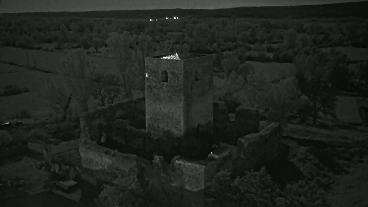 An aerial nighttime view shows the stone ruins of a tower and surrounding buildings, situated amidst dark trees and fields under a dark sky.