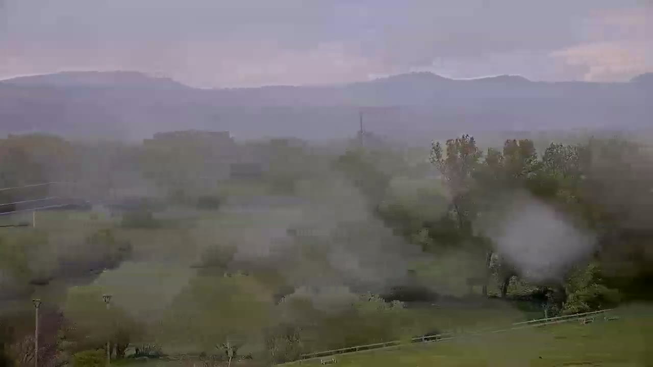 A hazy, rain-swept landscape of rolling hills, trees, and fields stretches toward distant mountains under a cloudy sky.