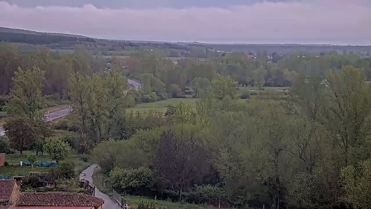 An elevated view of a verdant landscape under a cloudy sky shows a winding road traversing a tree-lined valley.