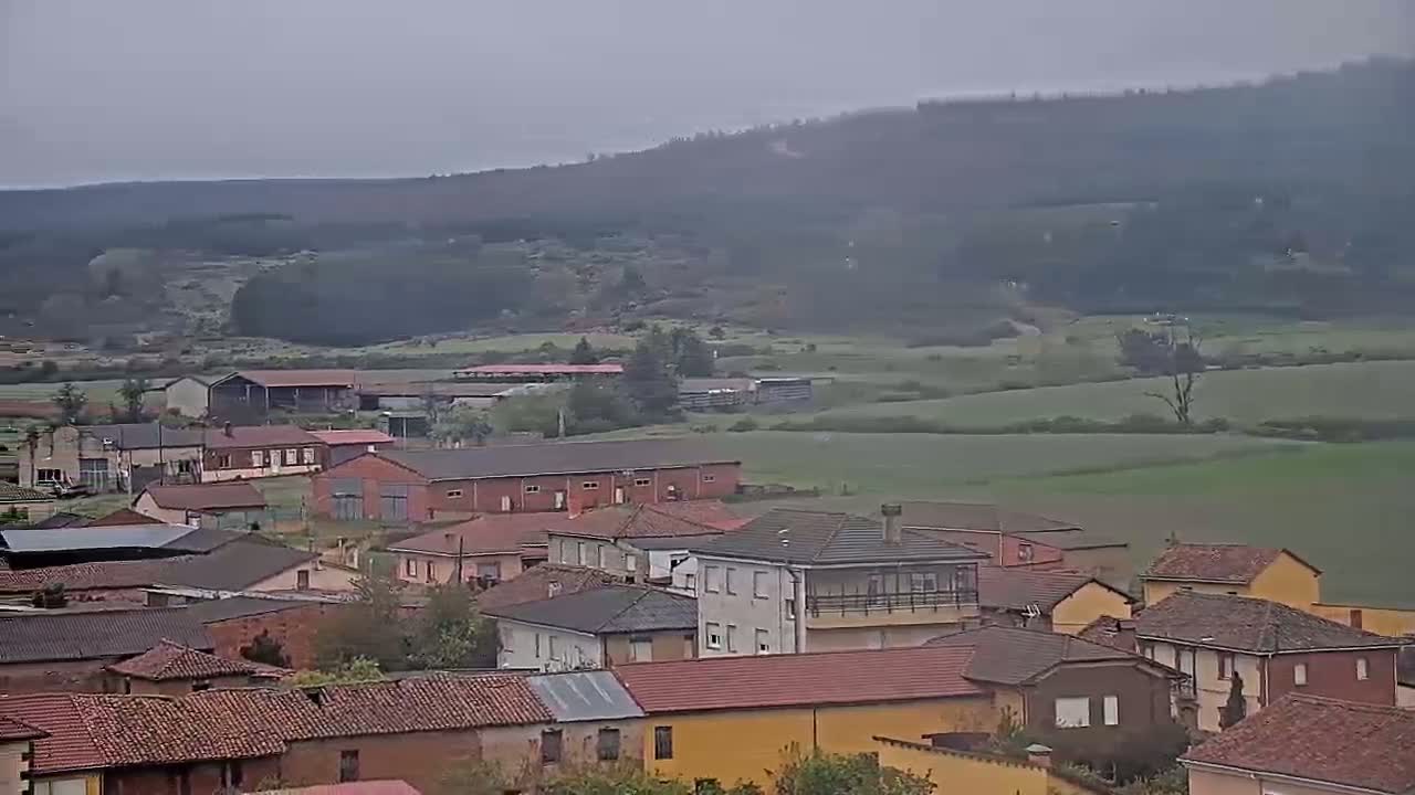 A small village with red-tiled roofs nestles in a valley under an overcast sky, with verdant fields and a distant, hazy hill in the background.